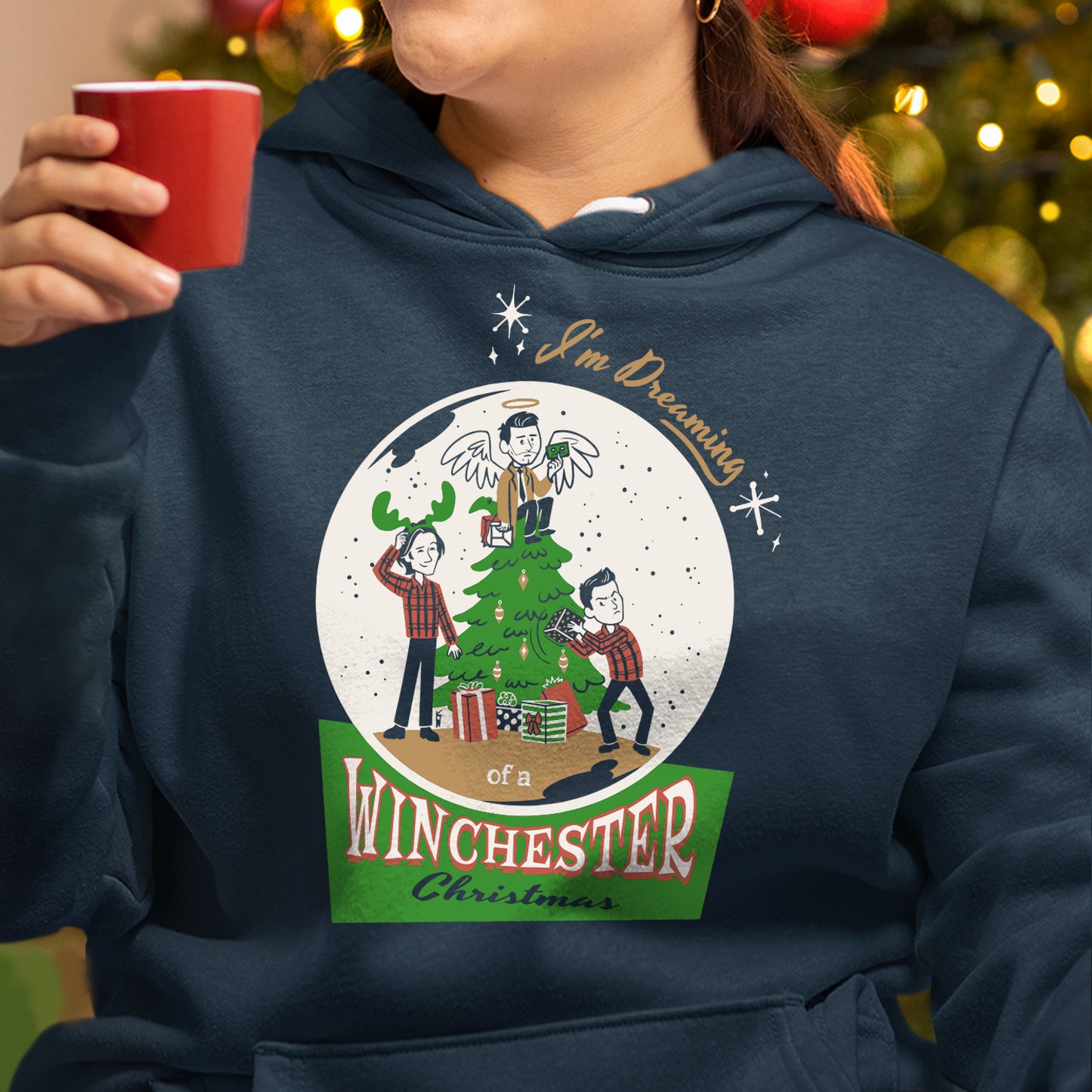 A brunette woman from the chin down, holding a red mug and wearing a dark blue pullover hoodie. On the front is a white globe with drawings of Dean Winchester, Sam Winchester, and Castiel, all working to put up a Christmas Tree. Text at the top and bottom says "I'm dreaming of a Winchester Christmas."