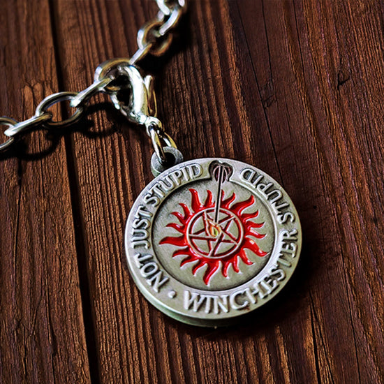 A flat circular charm that says "Not just stupid, Winchester stupid" around the edge, with an anti-possession symbol and a flaming arrow shot into the middle of it. Art by Charlotte Hill. Behind the charm is a wooden table.