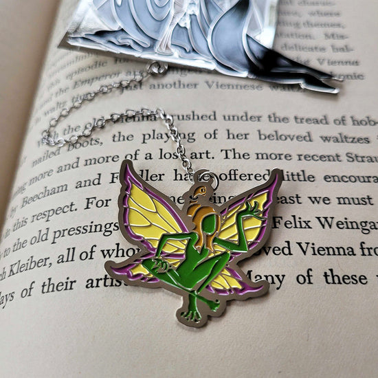 Close up of a charm attached to a bookmark, resting on the pages of a hardcover book. The charm depicts a green and yellow fairy.