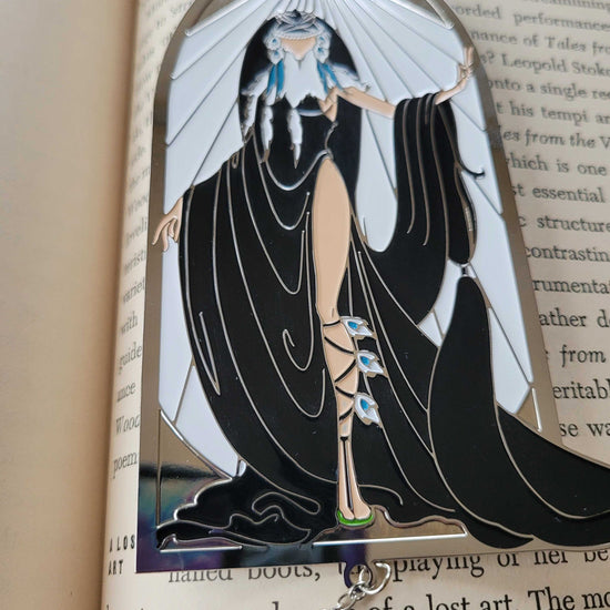 Close up view of a bookmark on the pages of a hardcover book. The bookmark depicts the ElfQuest character Winnowill, dressed in black robes with white rays above her