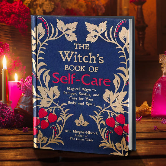 The cover of a hardcover copy of "The Witch's Book of Self-Care: Magical Ways to Pamper, Soothe, and Care for Your Body and Spirit". The cover is royal blue and gold with red strawberry accents.
