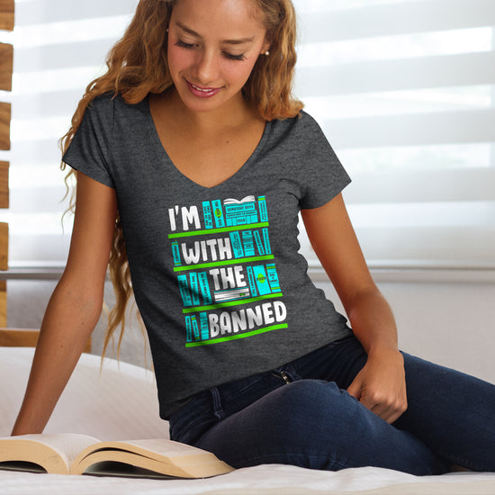 A female model wearing a grey T-shirt. There are four green bookshelves with light blue book spines covering each. The words "I'm With The Banned" are spread out across the four bookshelves in white text. The model is sitting on a white bed, reading a book. Behind her are white blinds covering a window.