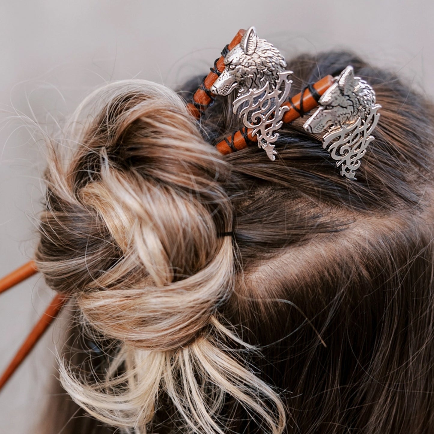 Top view of a long-haired model's head. Two wooden sticks are inserted through her hair, with silver wolfhead pendants at the end of each.