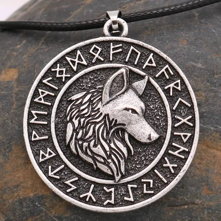 A close up view of a round stainless steel pendant, hanging from a black waxed rope. The pendant depicts the face of a wolf, shown in profile. Around the wolf face is the Norse Runic alphabet. Behind the pendant is a stone background, with gray and brown marbling.