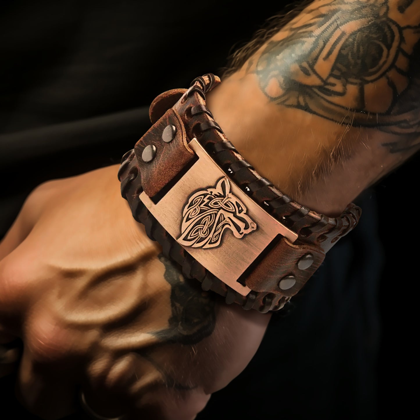 A picture of a man's tattooed arm wearing a brown leather cuff bracelet that features a copper plate with a wolf's head engraving on it.