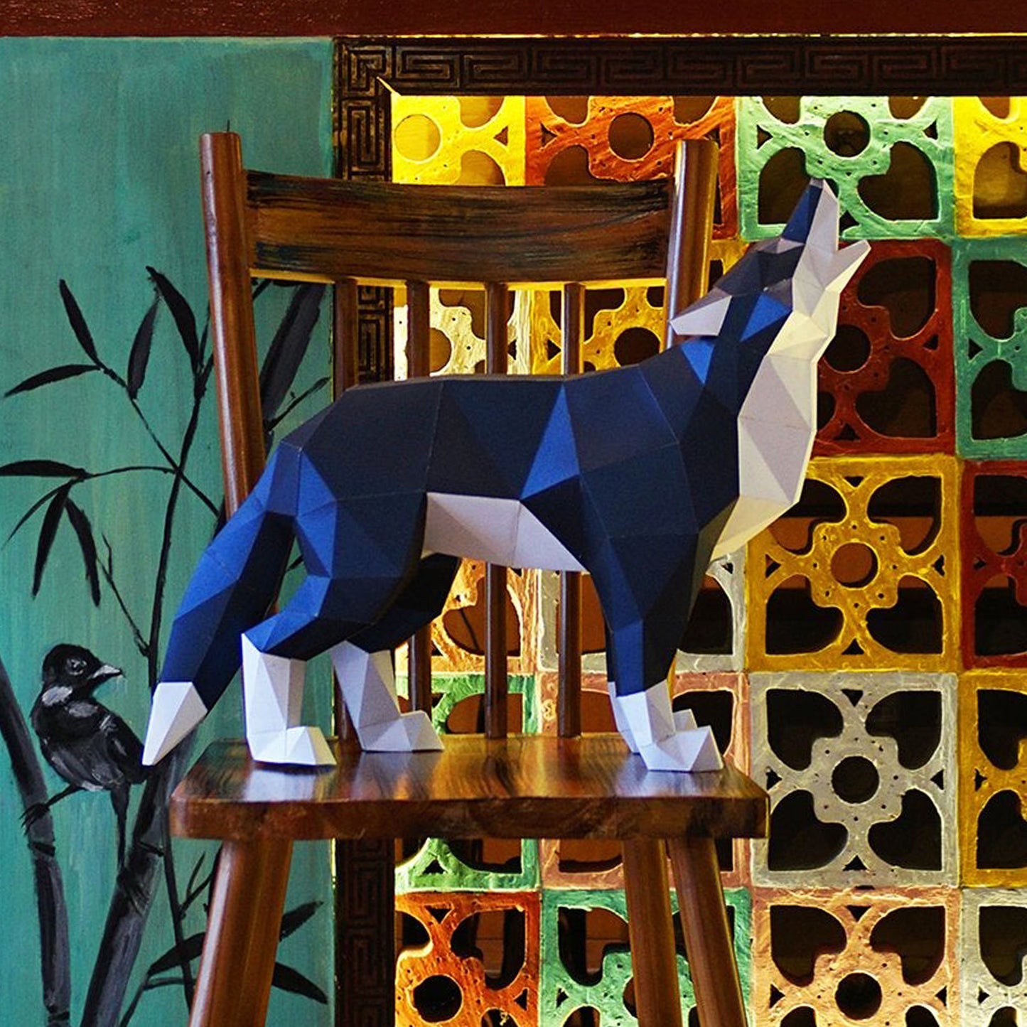 A blue and white paper model of a howling wolf, standing on a wooden stool. Behind the model is a multicolored wall with painted tree branches on one side.