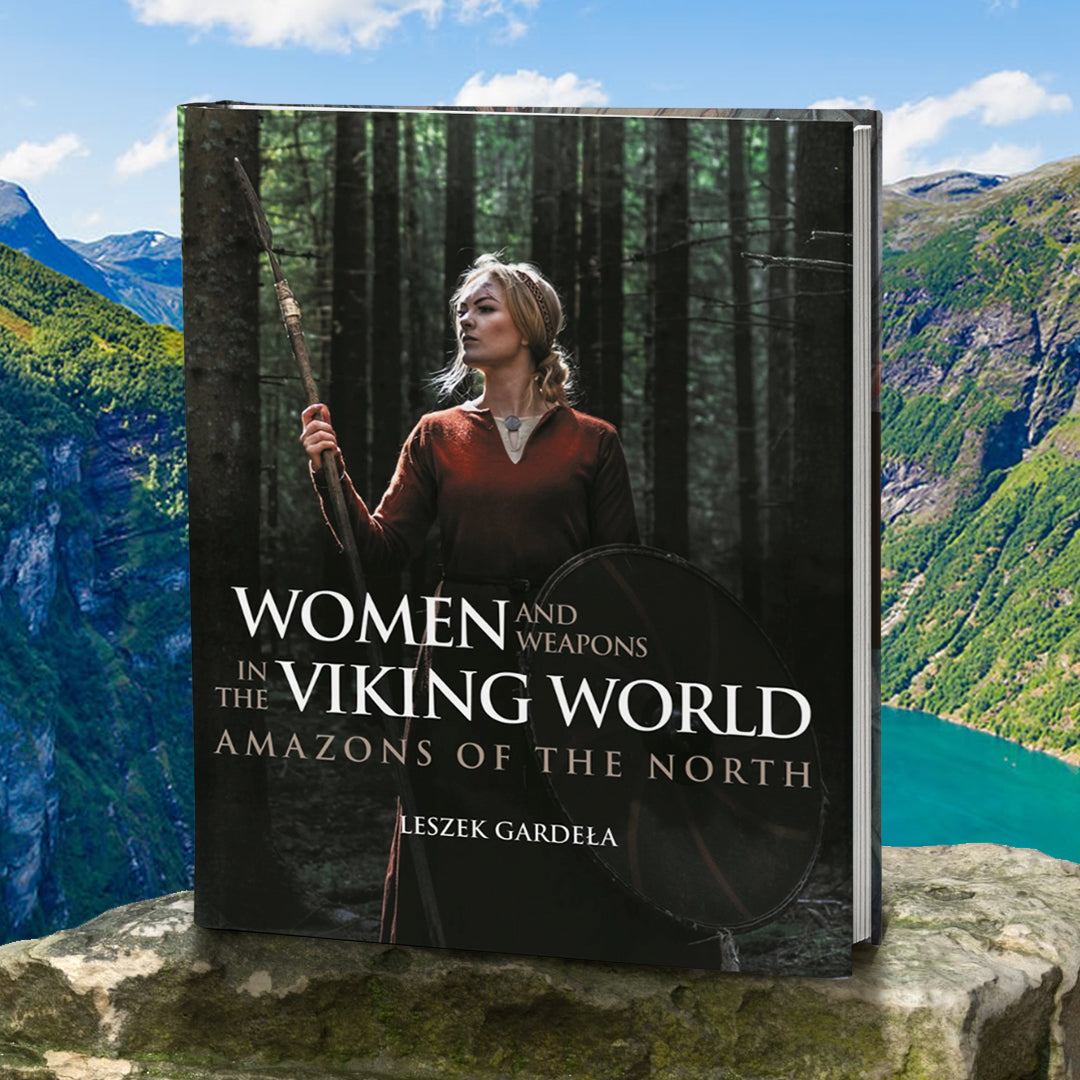A book cover sitting on a rock, with rolling hills and a river behind it. On the cover, a blonde woman in a red Viking outfit standing in a forest of trees. In one hand she is carrying a viking shield, in the other is a wooden spear. Below her is white text that reads “Women and weapons in the viking world: amazons of the north.”