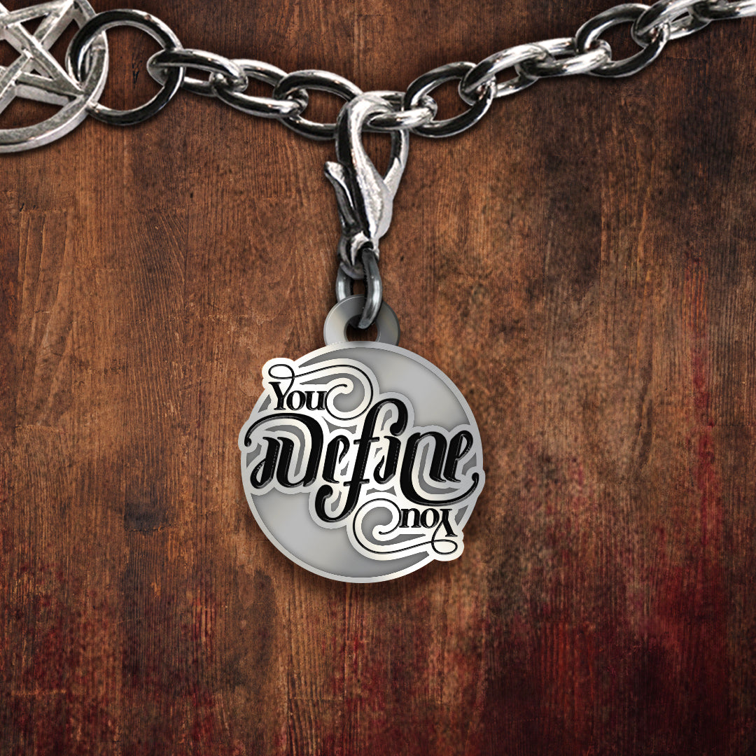 A silver-toned round pendant charm on a chain, featuring the phrase 'You define not' in black script with a white outline, displayed against a dark brown wood background.