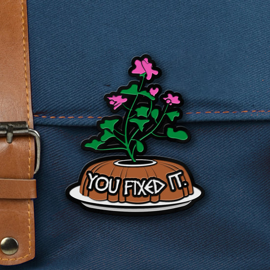 An enamel pin depicting a bundt cake with flowers and 'YOU FIXED IT' text on a blue background