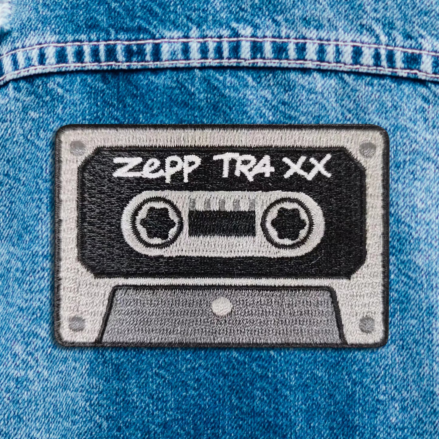 Close up of a black and grey iron on patch, depicting a cassette tape. On the front is white text saying "zepp traxx"