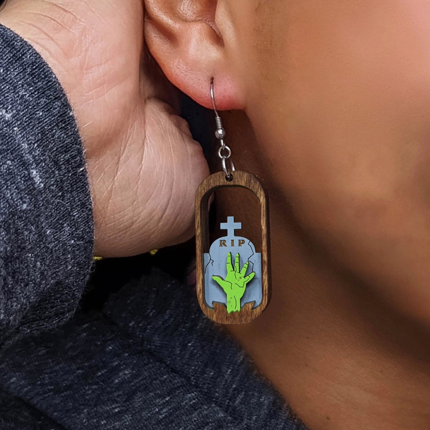A female model wearing an oval-shaped wooden earring. The center of the earring is open, with a green zombie hand reaching upward in front of a tombstone.