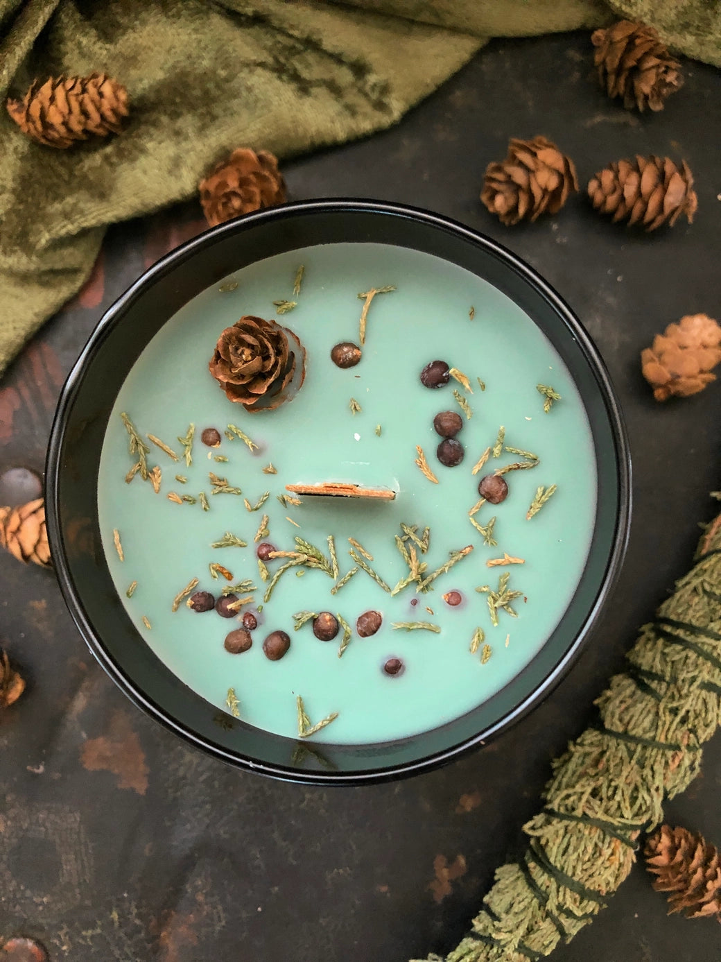 Candle with decorative elements on a dark surface with pine cones and green fabric.