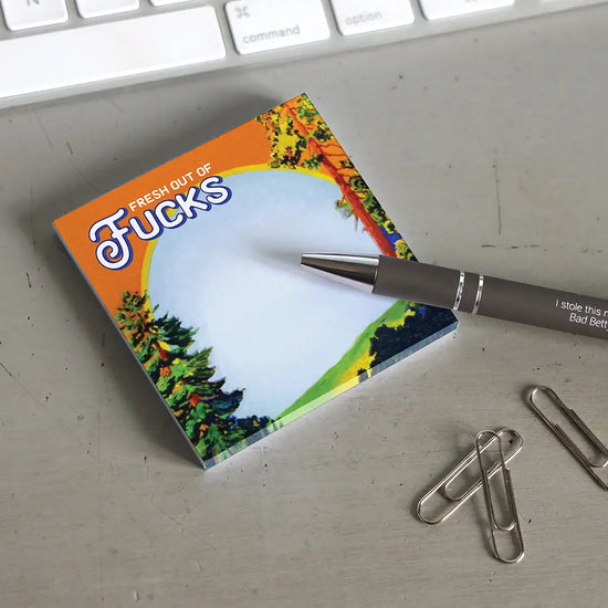 A sticky note on a white background. The sticky has a picture of trees in a meadown with a yellow arc at the top. White text says "fresh out of fucks." The note is on a grey desktop. Next to the note is a black pen, a handful of paperclips, and a computer keyboard
