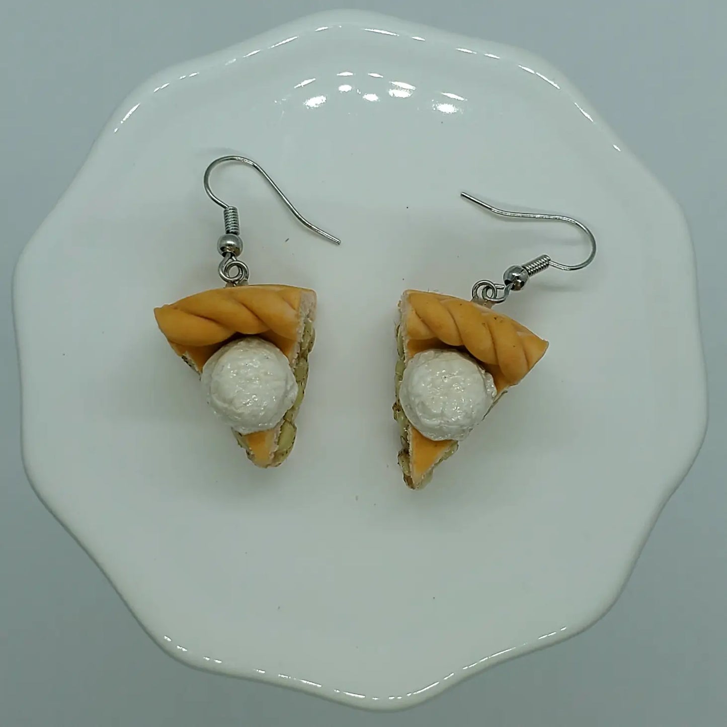 A pair of small, clay earrings in the shape of apple pie slices. You can see the "apple filling" from the side, and there is a scoop of ice cream on top. The earrings are on a set of silver earring hooks.