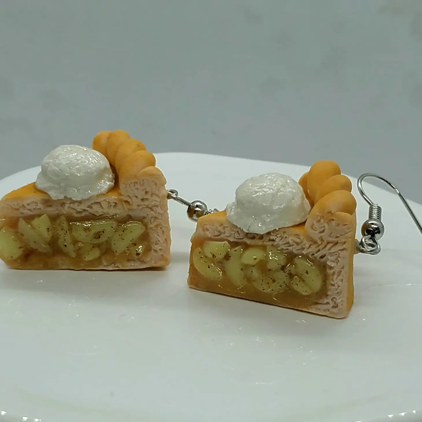 A pair of small, clay earrings in the shape of apple pie slices. You can see the "apple filling" from the side, and there is a scoop of ice cream on top. The earrings are on a set of silver earring hooks.