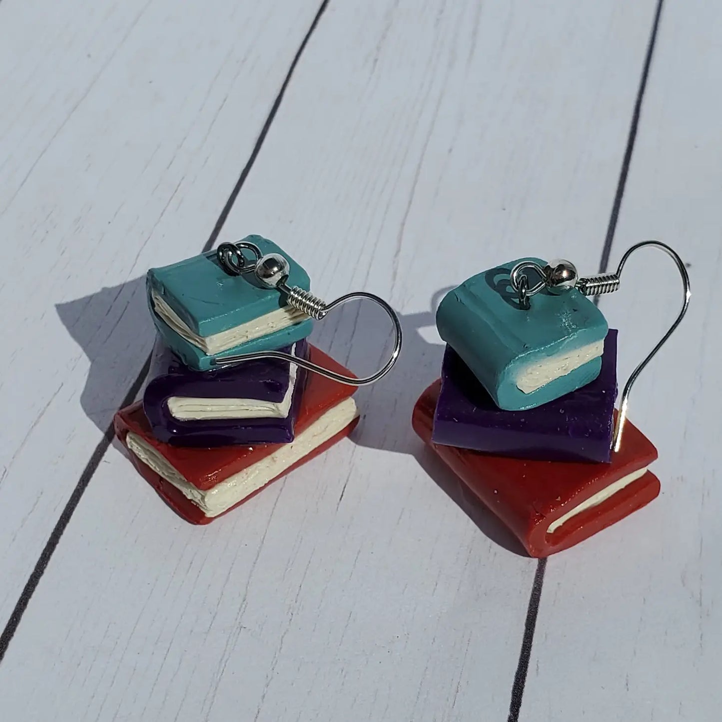 Load image into Gallery viewer, A pair of small, clay earrings in the shape of a stack of books, on a wooden table. The earrings are on a set of silver earring hooks. 

