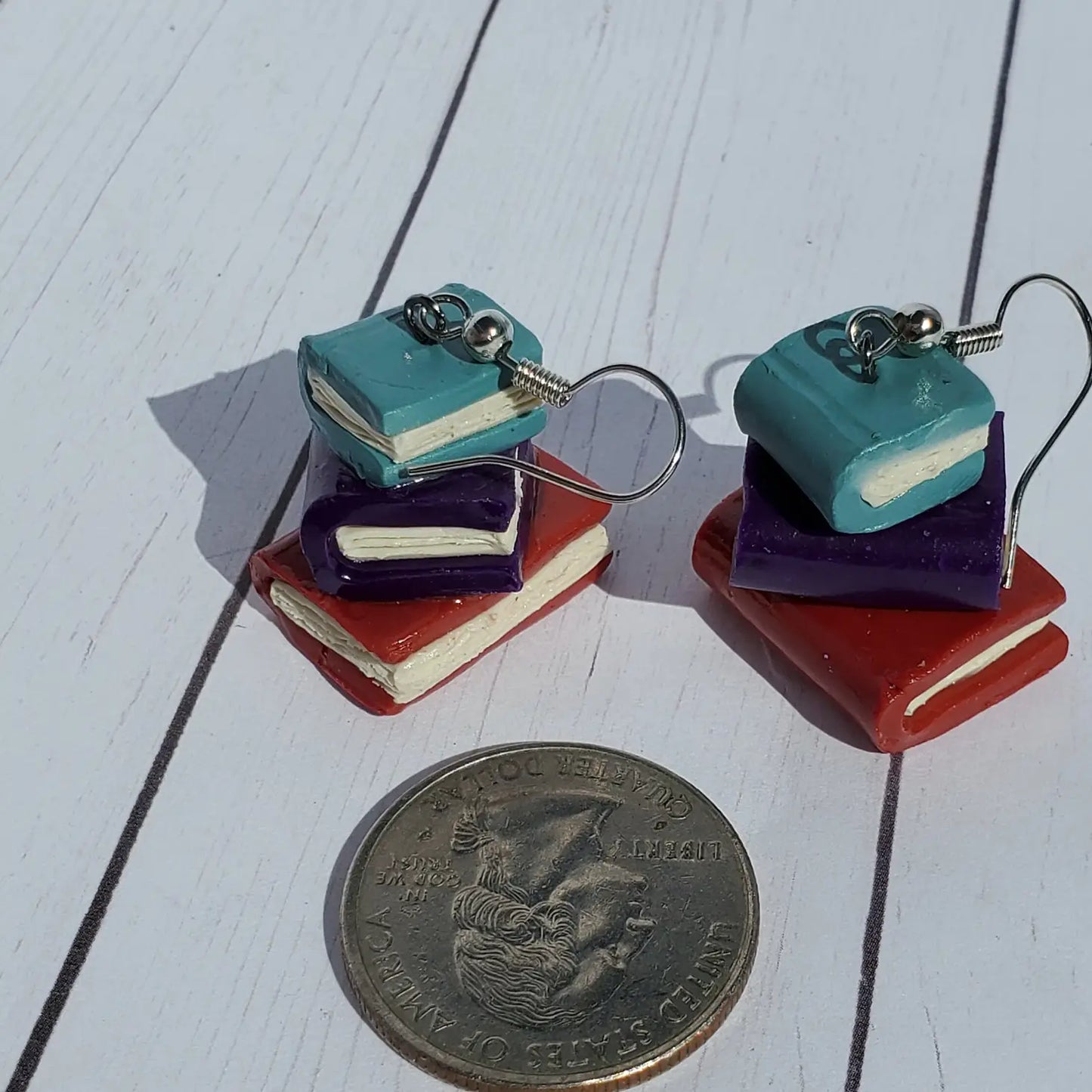 Load image into Gallery viewer, A pair of small, clay earrings in the shape of a stack of books, on a wooden table. The earrings are on a set of silver earring hooks. In front of the earrings is a US quarter, to show the size of the earrings in scale.
