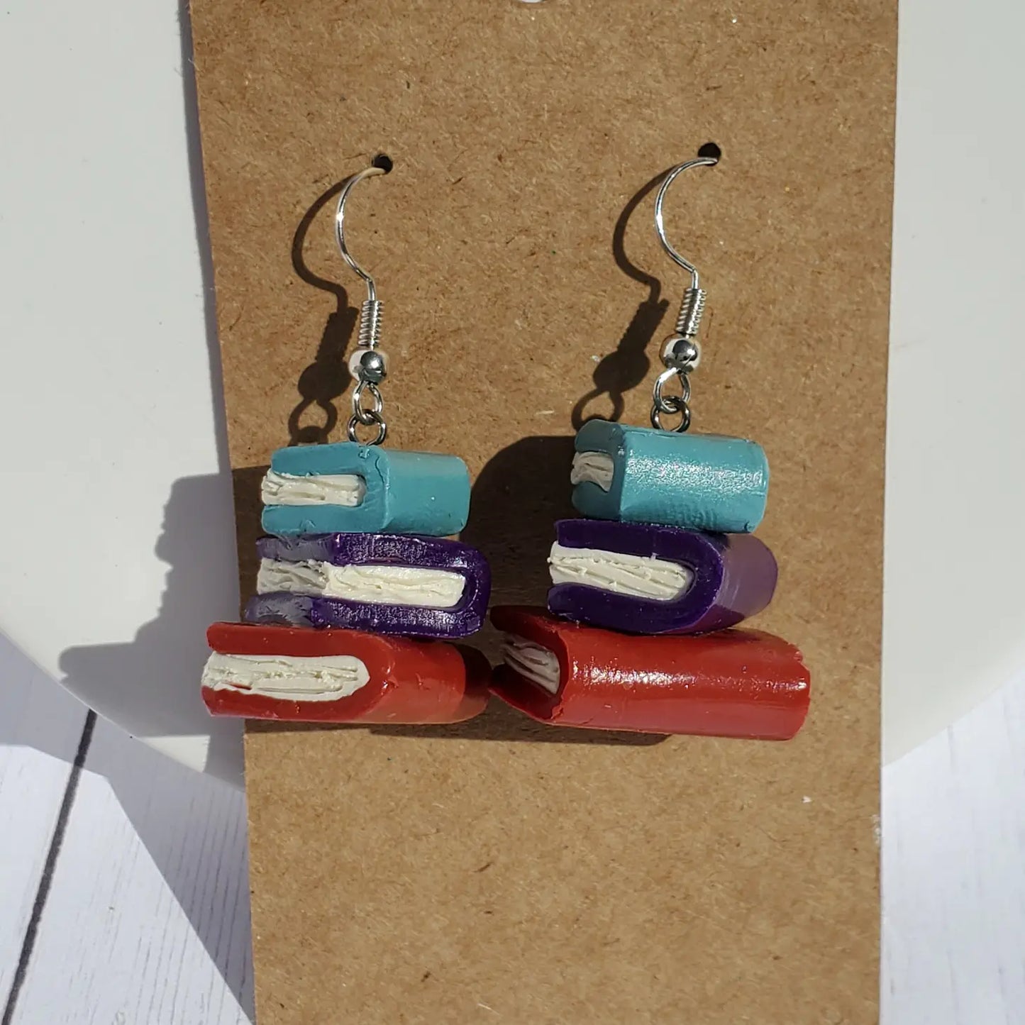 A pair of small, clay earrings in the shape of a stack of books hanging from a cardboard sheet, against a wooden table. The earrings are on a set of silver earring hooks. 