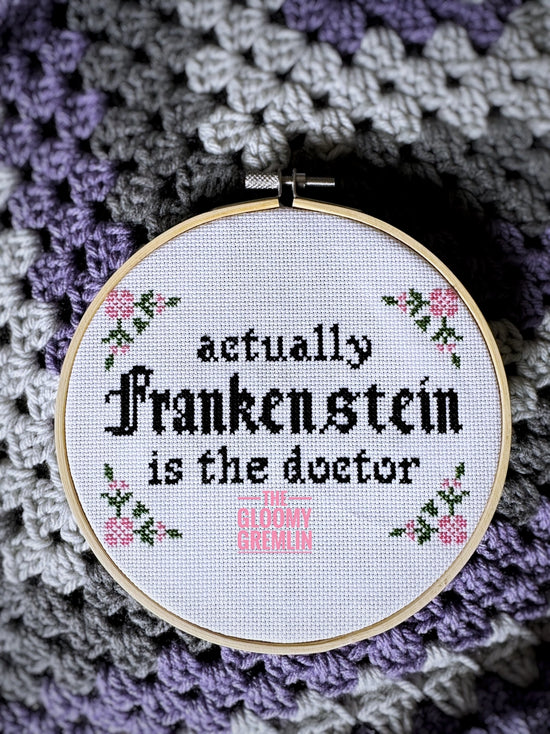 Embroidery hoop with cross-stitched text 'Actually Frankenstein is the doctor' on a crocheted background.