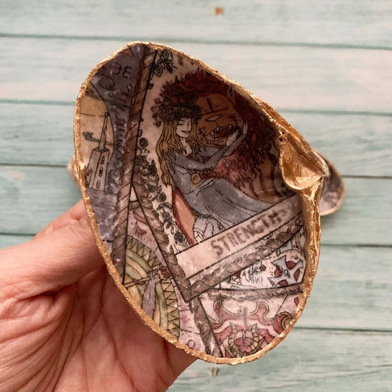 A seashell held in a model's hand, against a wooden background. The interior of the shell is decorated with tarot card drawings.