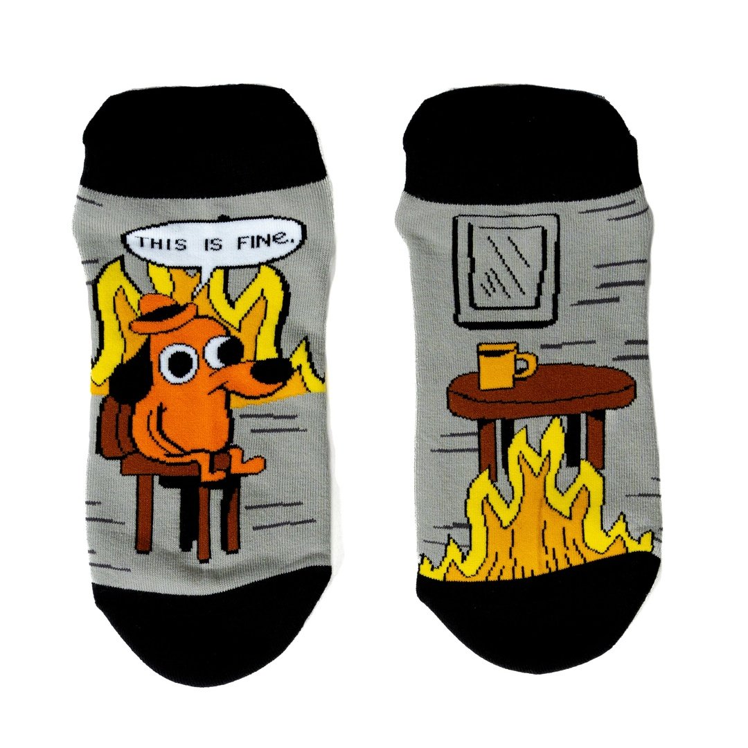 A pair of grey and back socks against a white background. One sock has a cartoon of a smilingbrown dog wearing a haat, sitting in a chair in front of burning flames. Above him is a caption saying "This is fine." The other sock has a brown wooden table with a coffee cup, and a roaring flame underneath it. The cuff and toes of the socks are black.