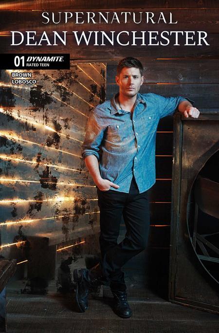 A comic book cover for 'Supernatural: Dean Winchester' issue 01, featuring a man in a blue denim shirt and black pants leaning against a rustic wooden wall with warm light streaks.