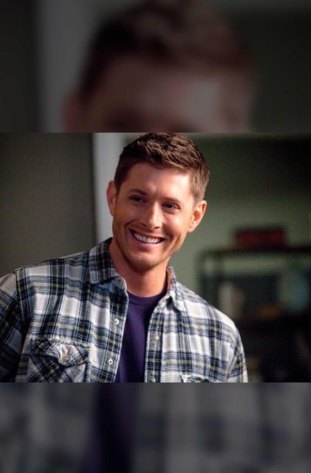 A smiling man with short brown hair wearing a blue and white plaid button-up shirt over a dark purple t-shirt, set against a blurred indoor background.