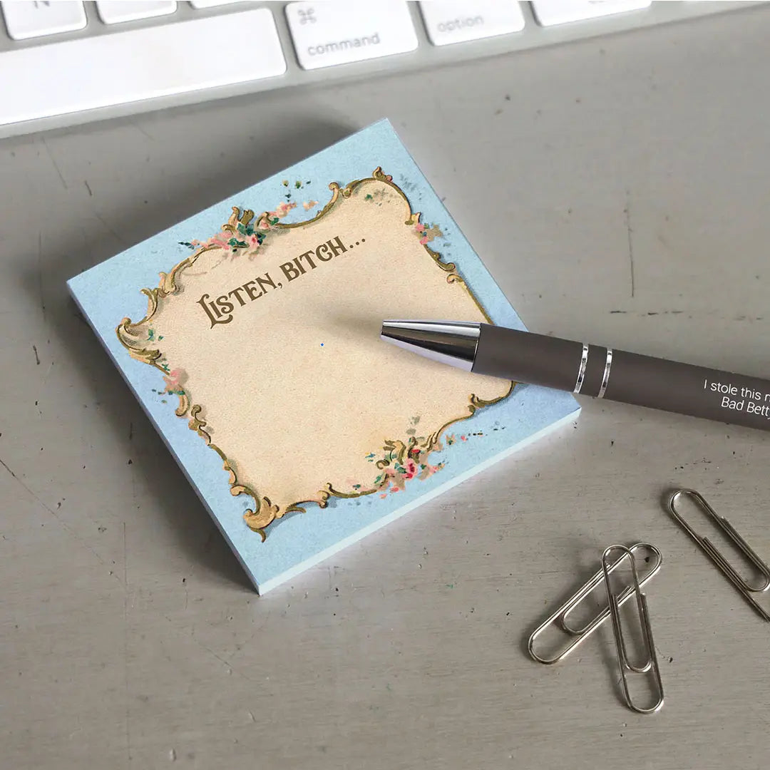 A blue sticky note with a parchment colored center, on a grey desktop. At the top is flowered filigree, with text saying "Listen, bitch." Next to the pad is a black pen, a handful of paperclips, and a computer keyboard