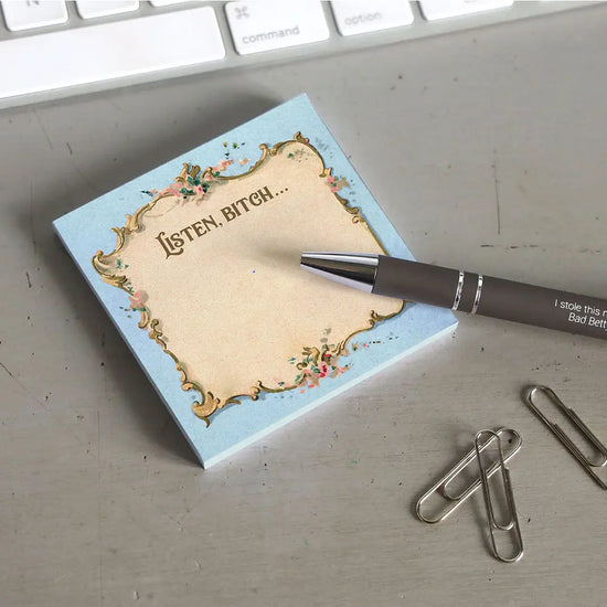 A blue sticky note with a parchment colored center, on a grey desktop. At the top is flowered filigree, with text saying "Listen, bitch." Next to the pad is a black pen, a handful of paperclips, and a computer keyboard