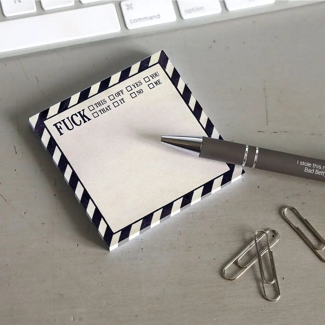 A white sticky note with black diagonal lines around the border. At the top is black text saying "fuck" with various checkable options next to it, including "this, off, you, and no". The note is on a grey desktop. Next to the note is a black pen, a handful of paperclips, and a computer keyboard