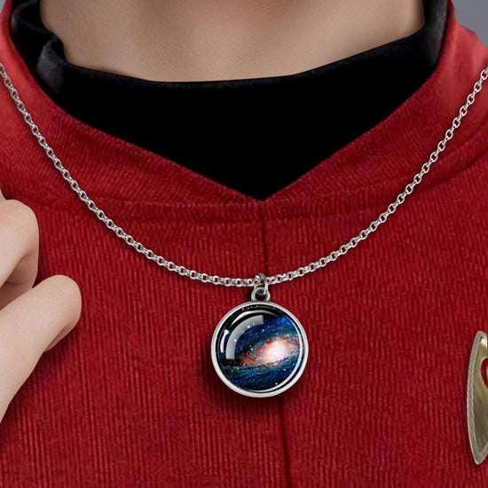 A silver circular necklace against a model’s red uniform shirt. The necklace features an image of a galaxy in space, under glass. 