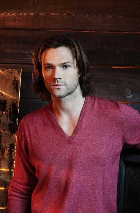 A man with shoulder-length brown hair wearing a heathered burgundy V-neck long-sleeve sweater, standing against a dark wood-paneled wall.