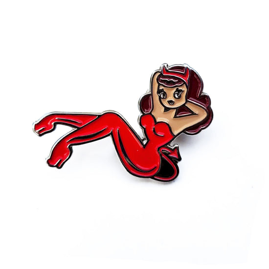 An enamel pin in the shape of a pin-up style woman lying back with her arms behind her head. She is wearing a red jumpsuit with devil horns and a devil's tail.