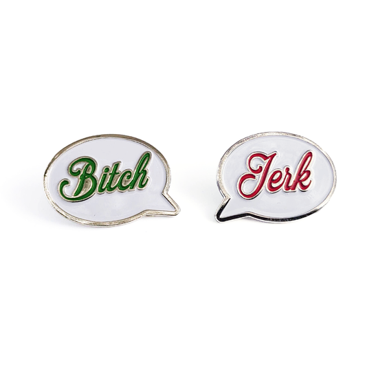 A set of two white enamel pins in the shape of speech bubbles. One reads "Bitch" in green script font. The other reads "Jerk" in red script font. 