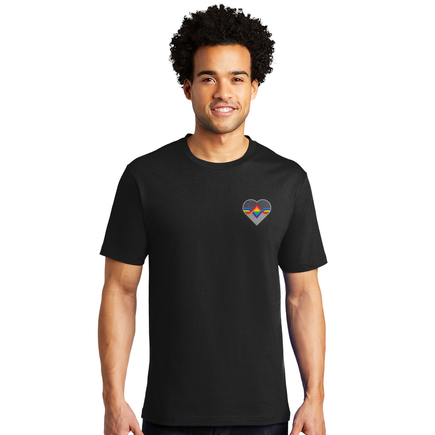 A male model wearing a black T-shirt. The Acting Ensign Pride symbol is at the top left corner of the shirt.  The symbol is a grey heart with rainbow lines running through the center.
