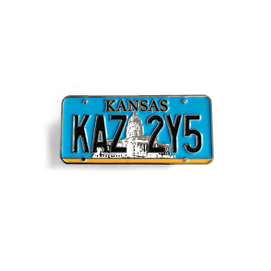 A miniature Kansas license plate against a white background. The pate reads KAZ 2Y5, with a white farmhouse behind the letters against a light blue background.